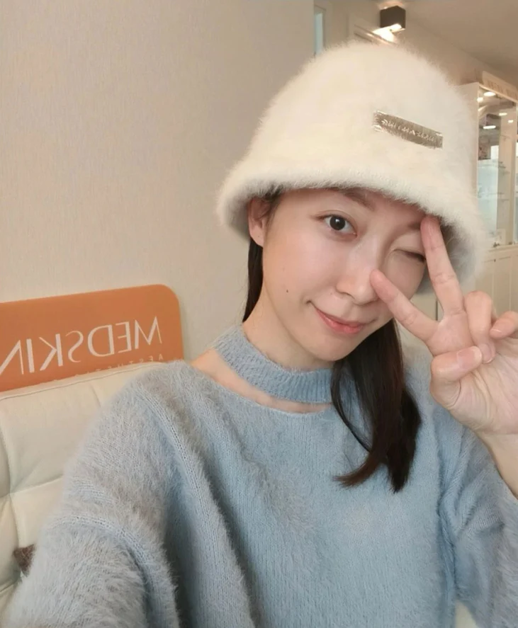 穿淺藍色針織上衣、戴白色毛絨漁夫帽的女士在室內比出勝利手勢自拍，面帶微笑，背景為診所環境