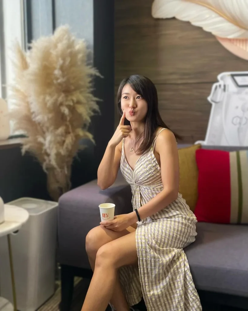 長髮女士身穿格紋吊帶連身裙，坐在灰色梳化上，右手食指輕觸嘴唇，左手拿着白色紙杯，身旁有乾花與紅色咕𠱸