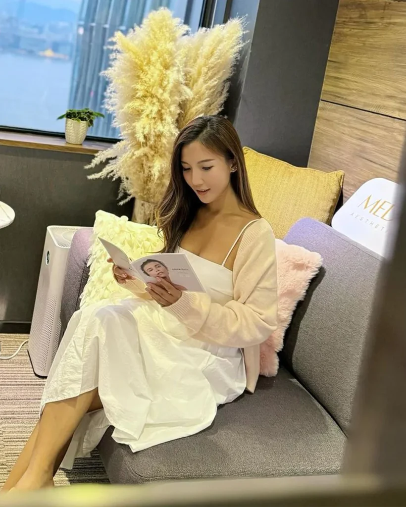 一名長髮女士穿白色吊帶裙配粉紅色外套，坐在灰色梳化上翻閱手上的療程小冊子，身旁有金色與粉紅色咕𠱸及乾花裝飾，背景為海景窗戶