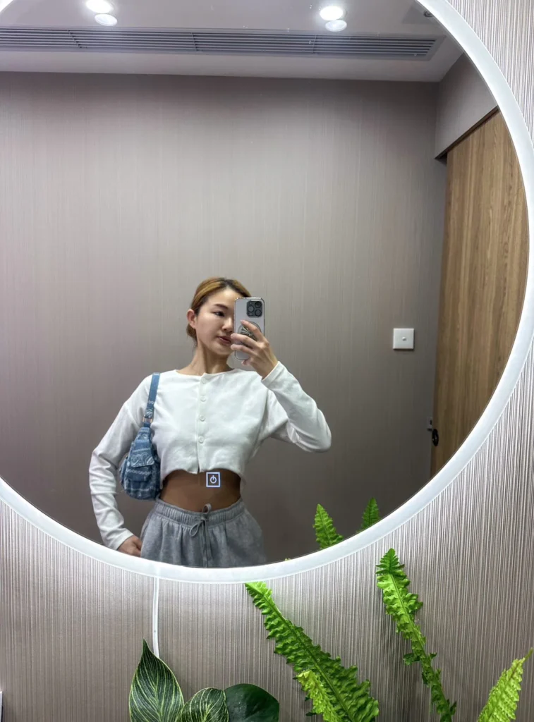 一名女子在圓形大鏡前用手機自拍，身穿白色長袖上衣及灰色運動褲，斜背藍色牛仔布小袋，鏡前下方擺放綠色盆栽。