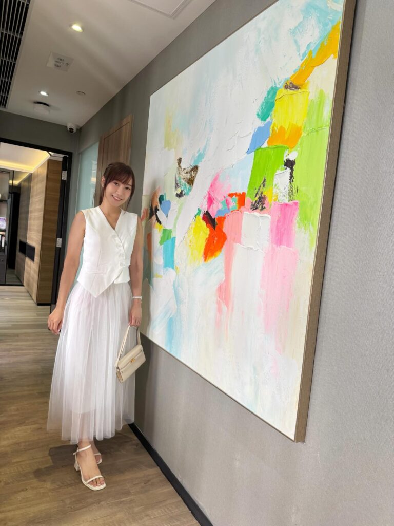 一位穿白色無袖上衣與白色長紗裙的女士，手持米白色小手袋，站在走廊一幅大型彩色抽象畫旁微笑拍照。