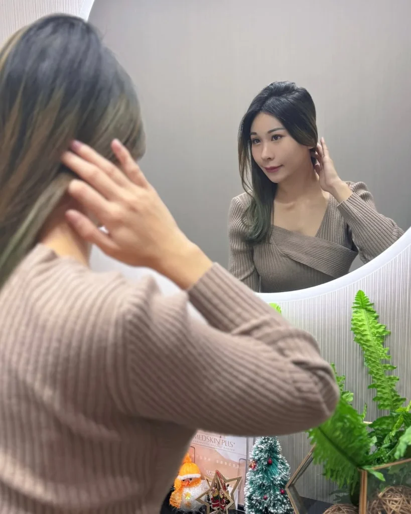 女士以手機對鏡自拍，身穿淺啡色羅紋上衣，鏡中映出本人身影，旁邊有綠色盆栽、聖誕擺設及診所標示牌