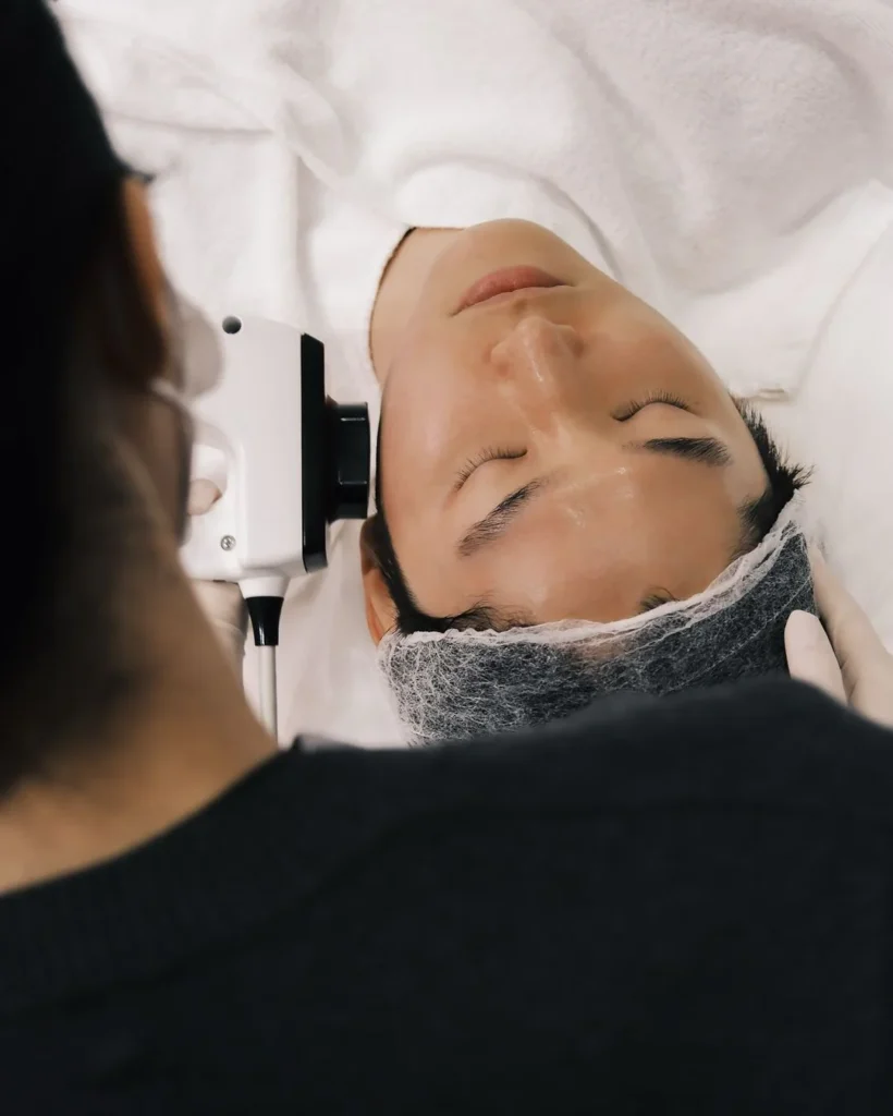 美容師戴手套手持白色儀器探頭，在包著紗布的女士額頭位置操作，女士閉眼躺於治療床上