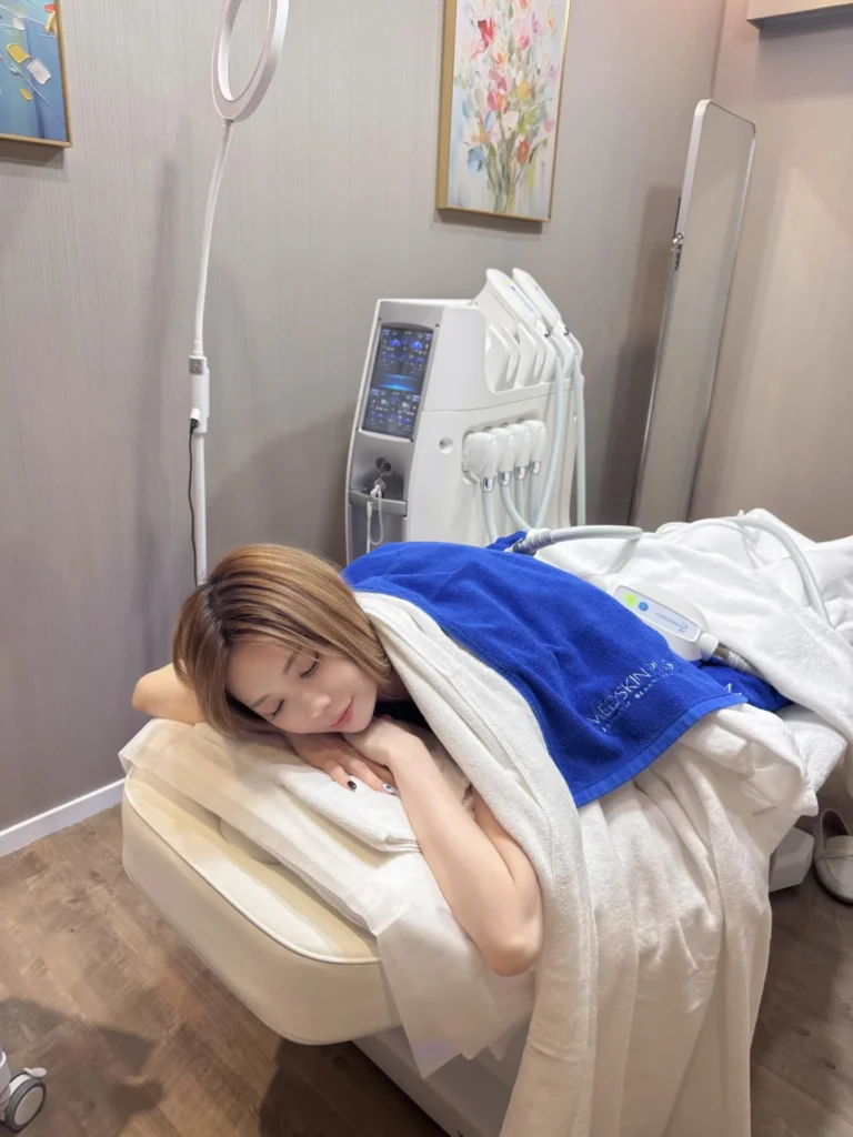 一名棕色長髮女士側躺在療程床上閉目休息，身上覆蓋白色毛巾及印有MEDSKIN PLUS+字樣的藍色毛巾，背後放置一部白色多功能美容儀器