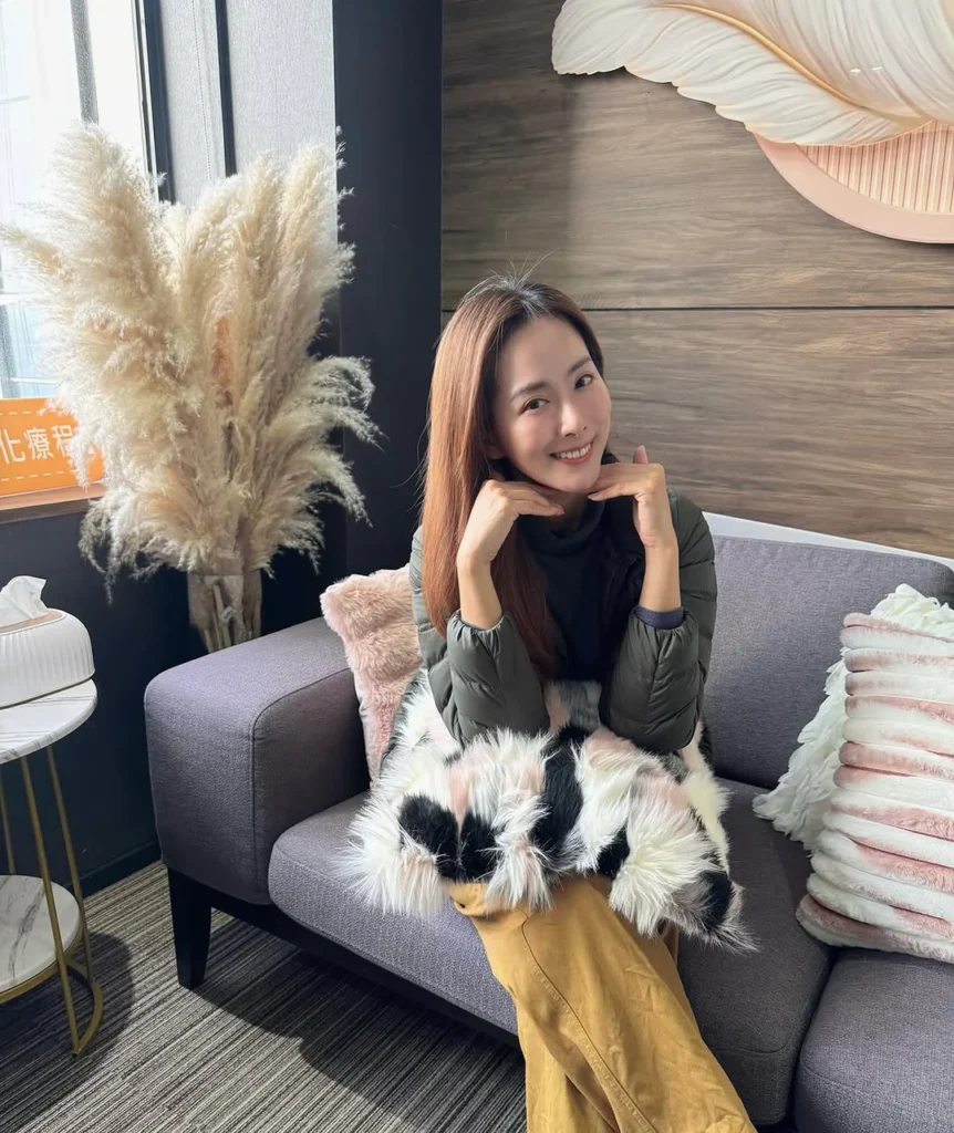 一位留長髮的女士坐在灰色梳化上，雙手輕托面頰微笑，身旁放置黑白毛絨咕𠱸及粉紅色毛毛咕𠱸。