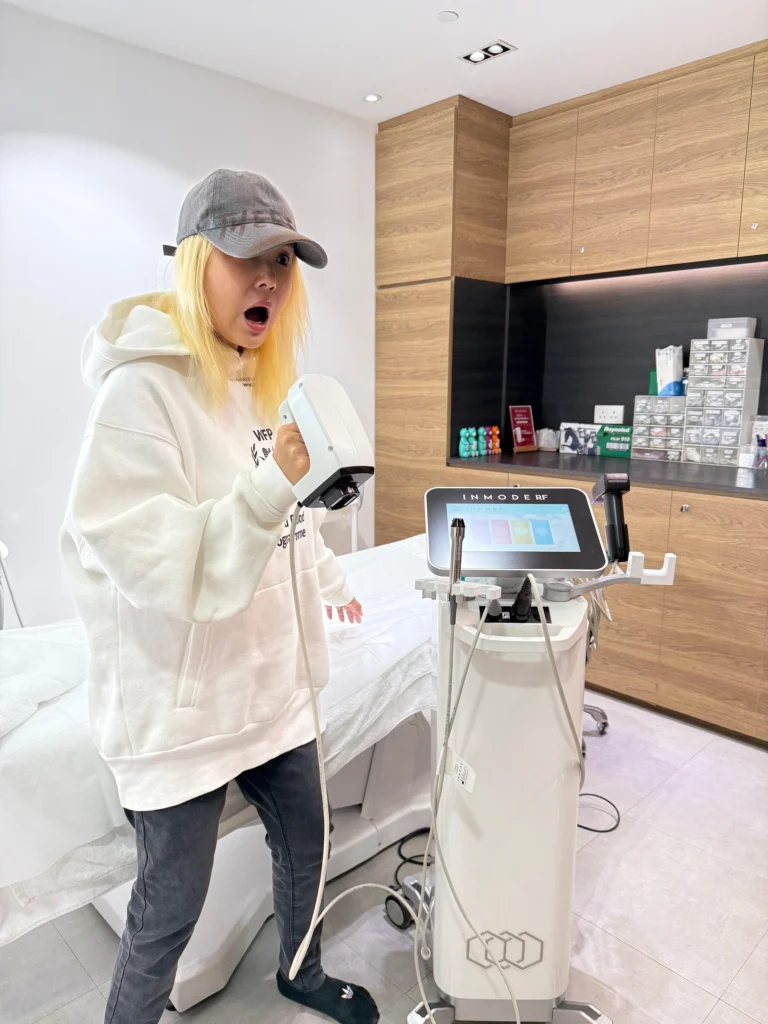 一名金髮女士戴灰色鴨舌帽、穿白色衛衣，在診所內手持白色的射頻探頭，身旁為 INMODE RF 儀器、觸控螢幕及治療床。