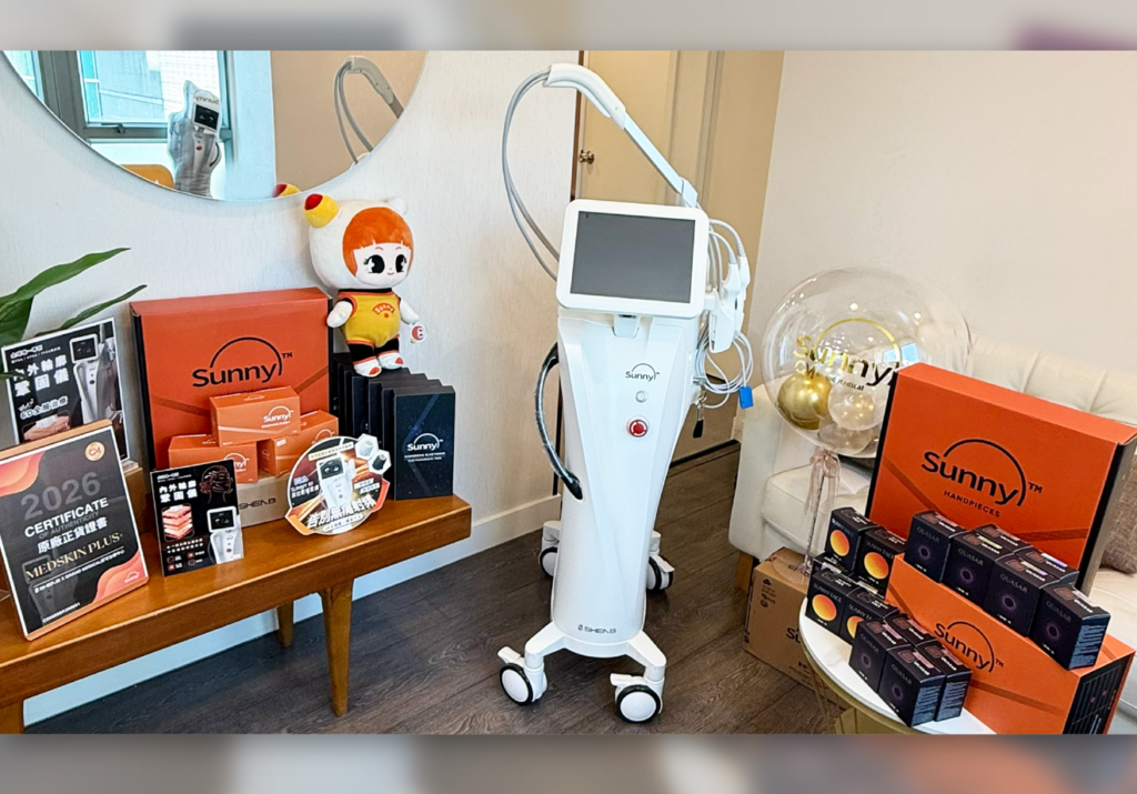 診所房間內擺放白色 SUNNY RF 射頻儀器，旁邊陳列橙色 SUNNY 包裝盒、黑色 HAND PIECES 探頭盒、原廠正貨認證展示牌及吉祥物公仔。